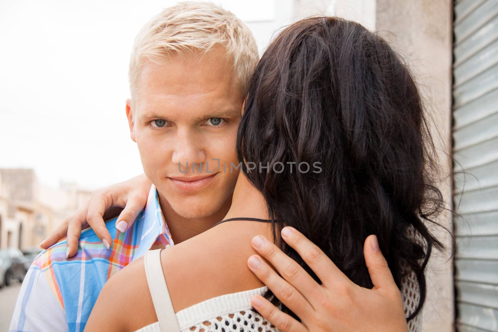 Romantic handsome young blond man hugging his girlfriend looking past the side of her head at the camera with a smile