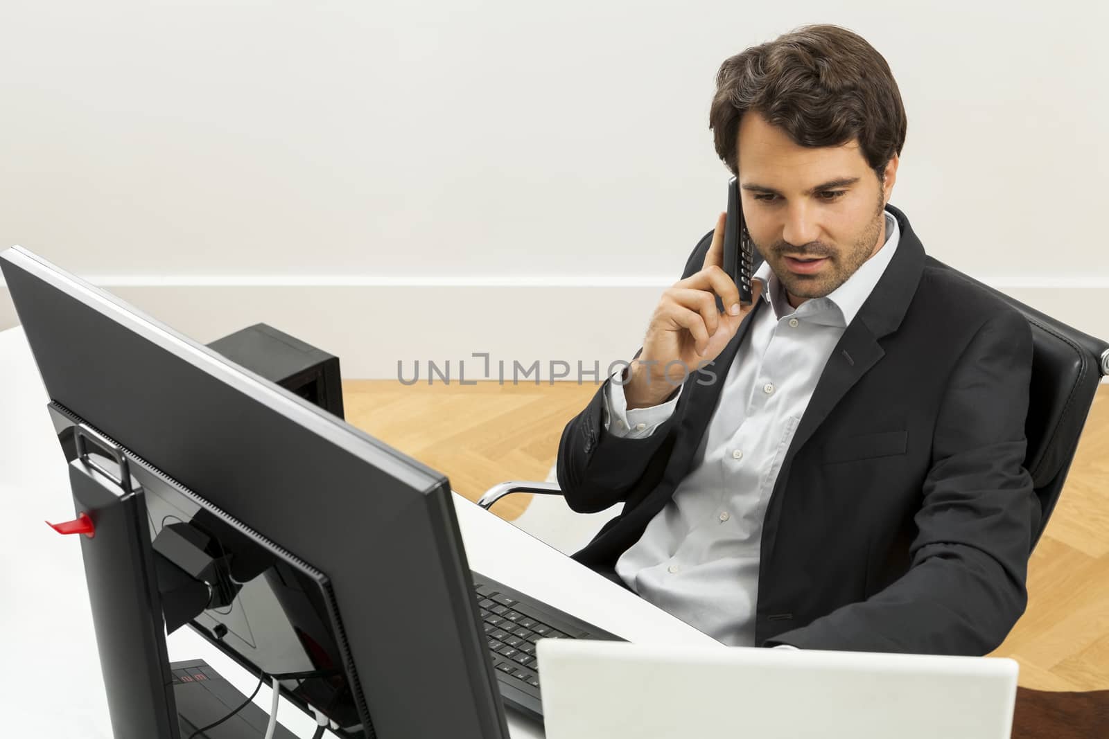 Successful businessman working in his office sitting in a chair at his desk and desktop computer listening to a call on his mobile phone, high angle side view