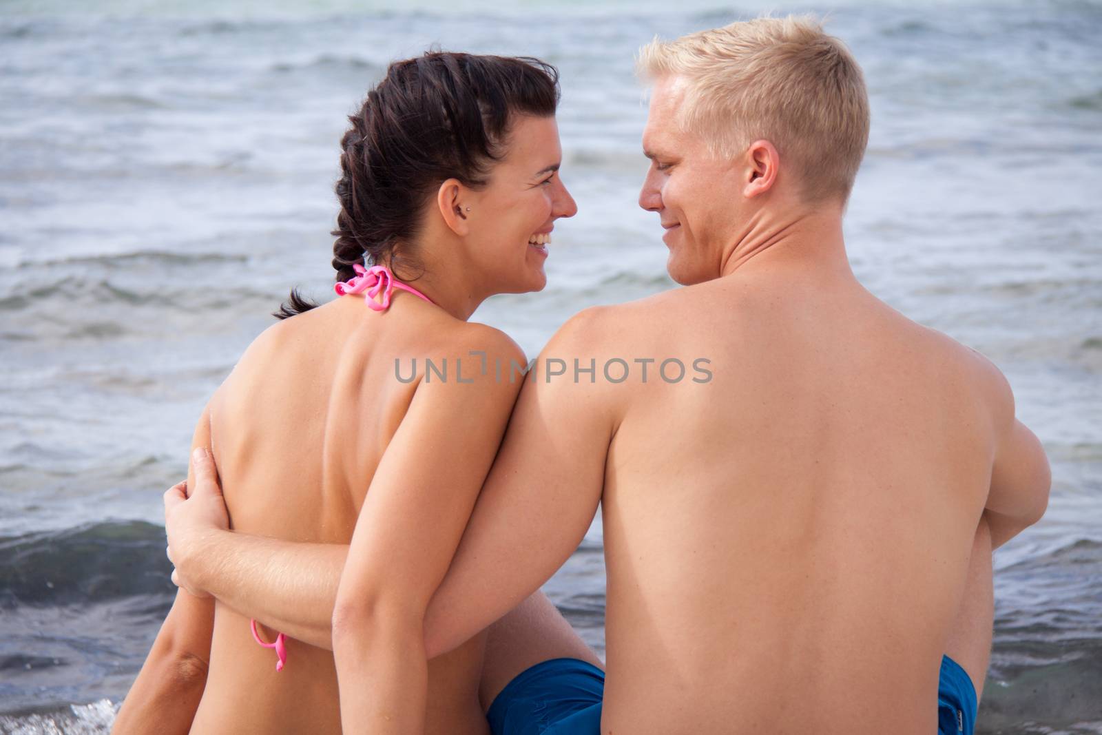 Happy romantic young couple enjoying a date at the seaside sitting arm in arm overlooking the ocean and looking back at the camera with charming happy smiles