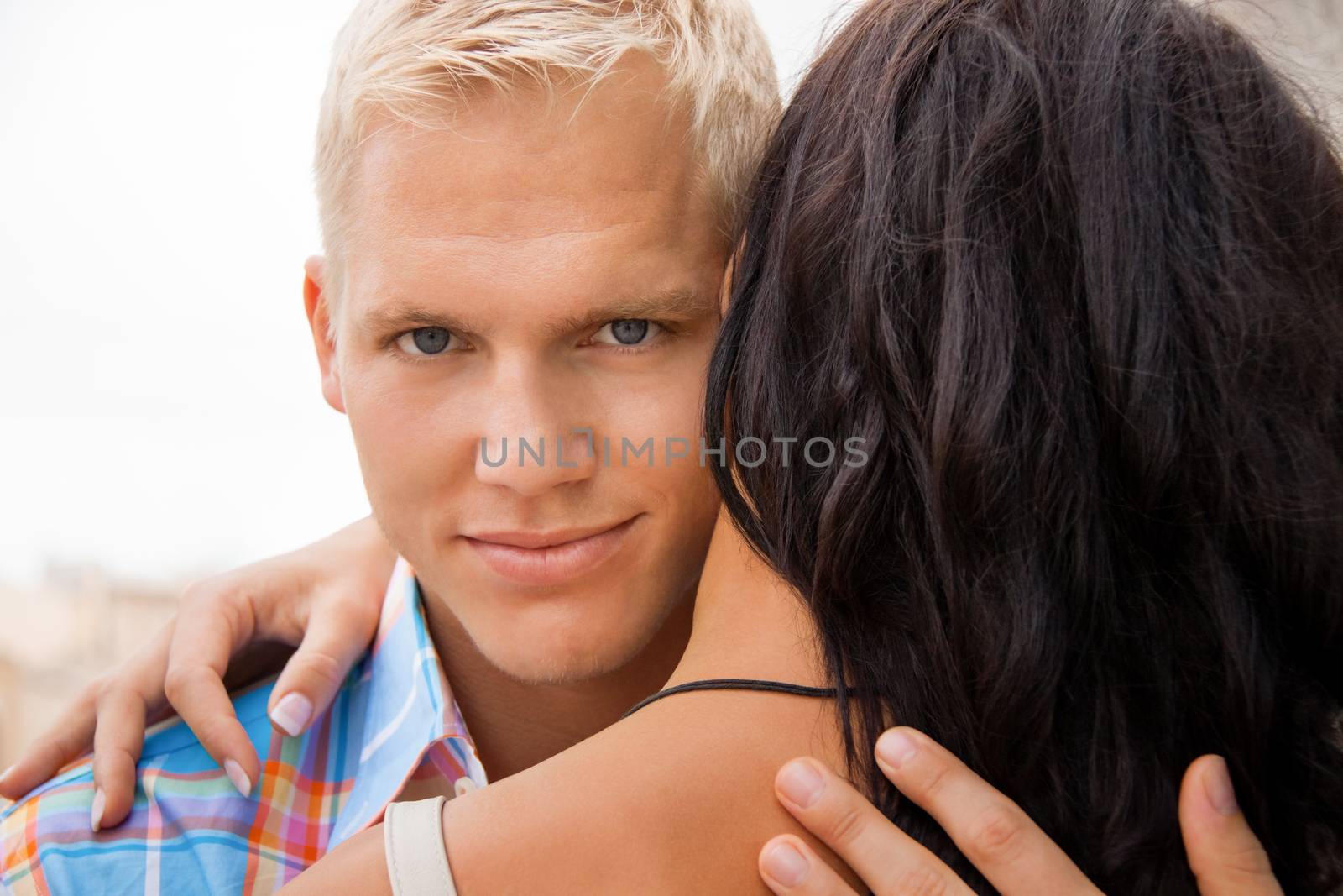 Romantic handsome young blond man hugging his girlfriend looking past the side of her head at the camera with a smile