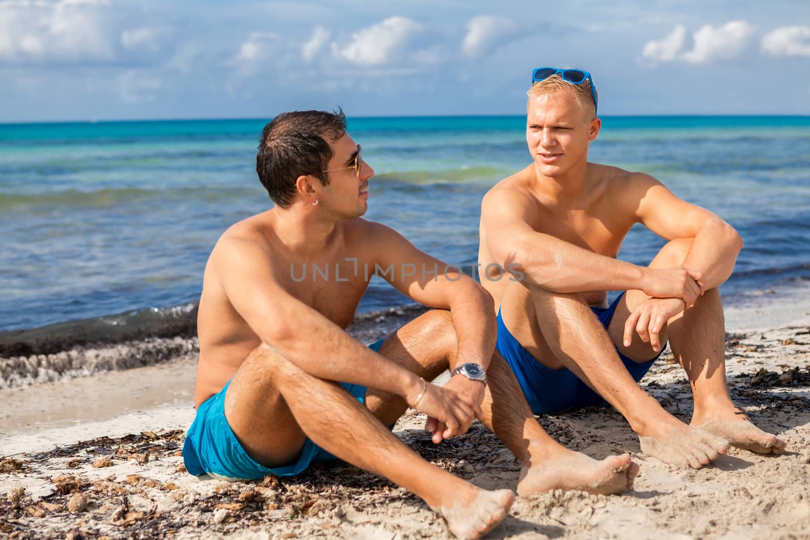 Two handsome young men chatting on a beach in their swimsuits sitting side by side on the sand with their backs to the ocean enjoying a relaxing summer day at the beach