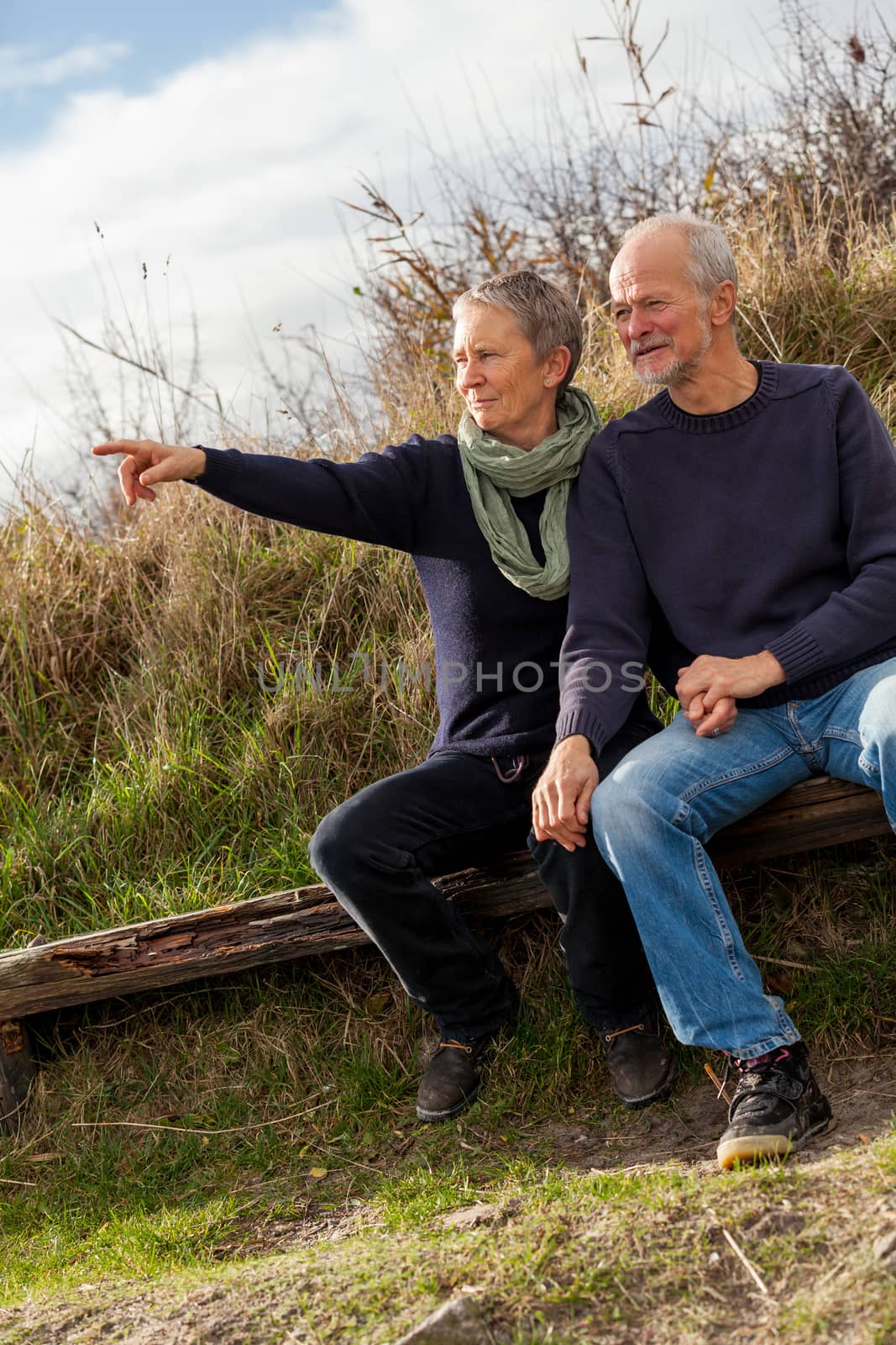 happy senior couple relaxing together in the sunshine on a wooden bench in the countryside with the one reclining full length on the seat with his head on his partners lap