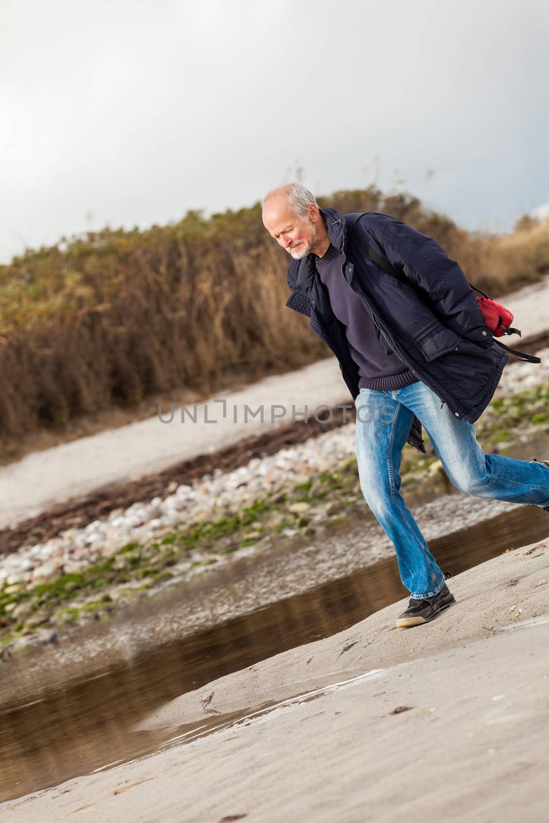 Elderly energetic man running along a beach at the edge of a bay on a cold day as he exercises to remain fit and healthy while enjoying nature