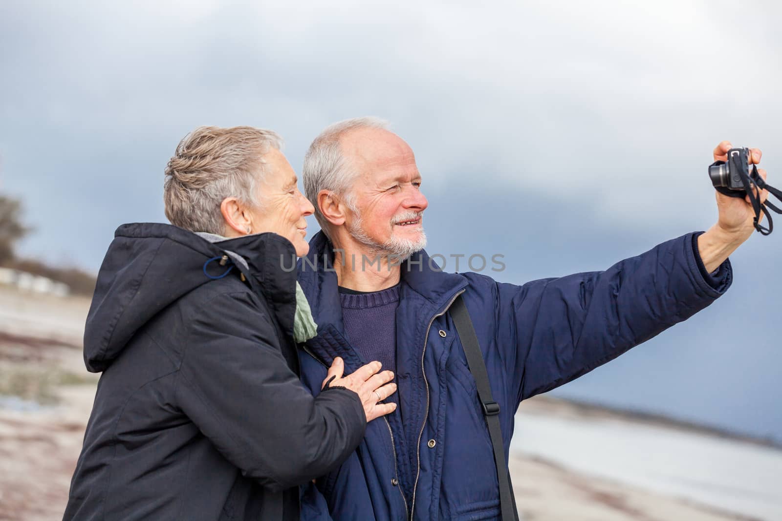 Elderly couple taking a self portrait on a compact digital camera posing in the open air and sunshine with their heads close together smiling at the lens