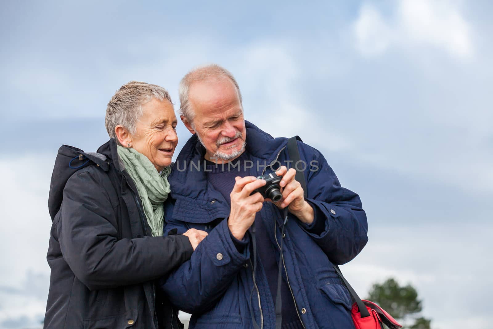 Elderly couple taking a self portrait on a compact digital camera posing in the open air and sunshine with their heads close together smiling at the lens