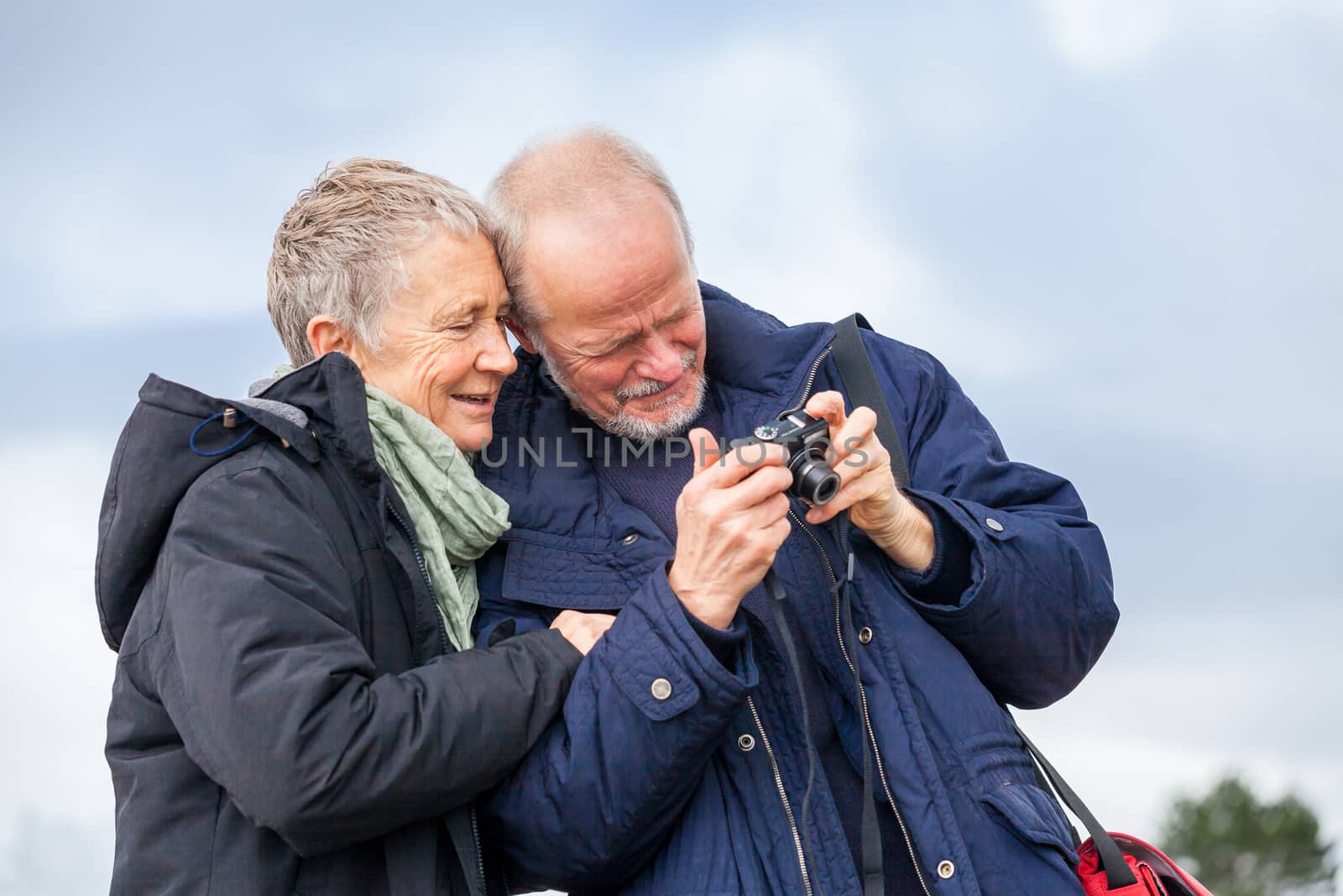 Elderly couple taking a self portrait on a compact digital camera posing in the open air and sunshine with their heads close together smiling at the lens