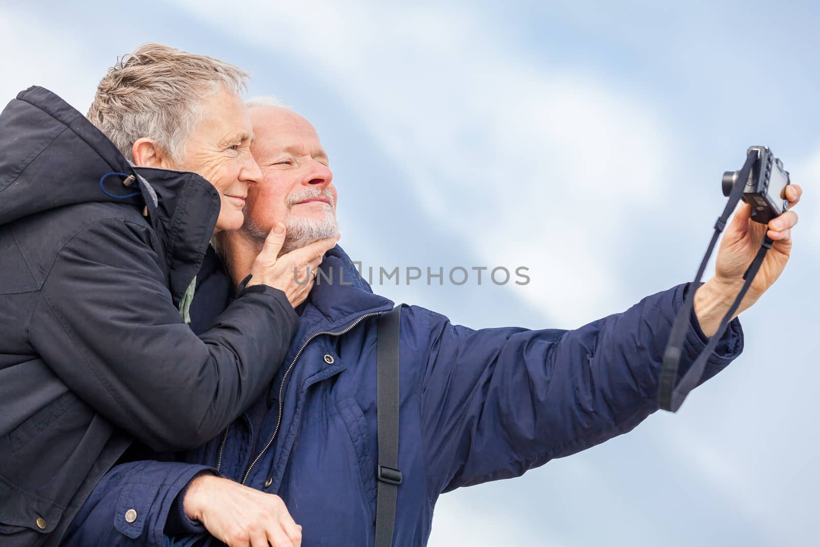 Elderly couple taking a self portrait on a compact digital camera posing in the open air and sunshine with their heads close together smiling at the lens