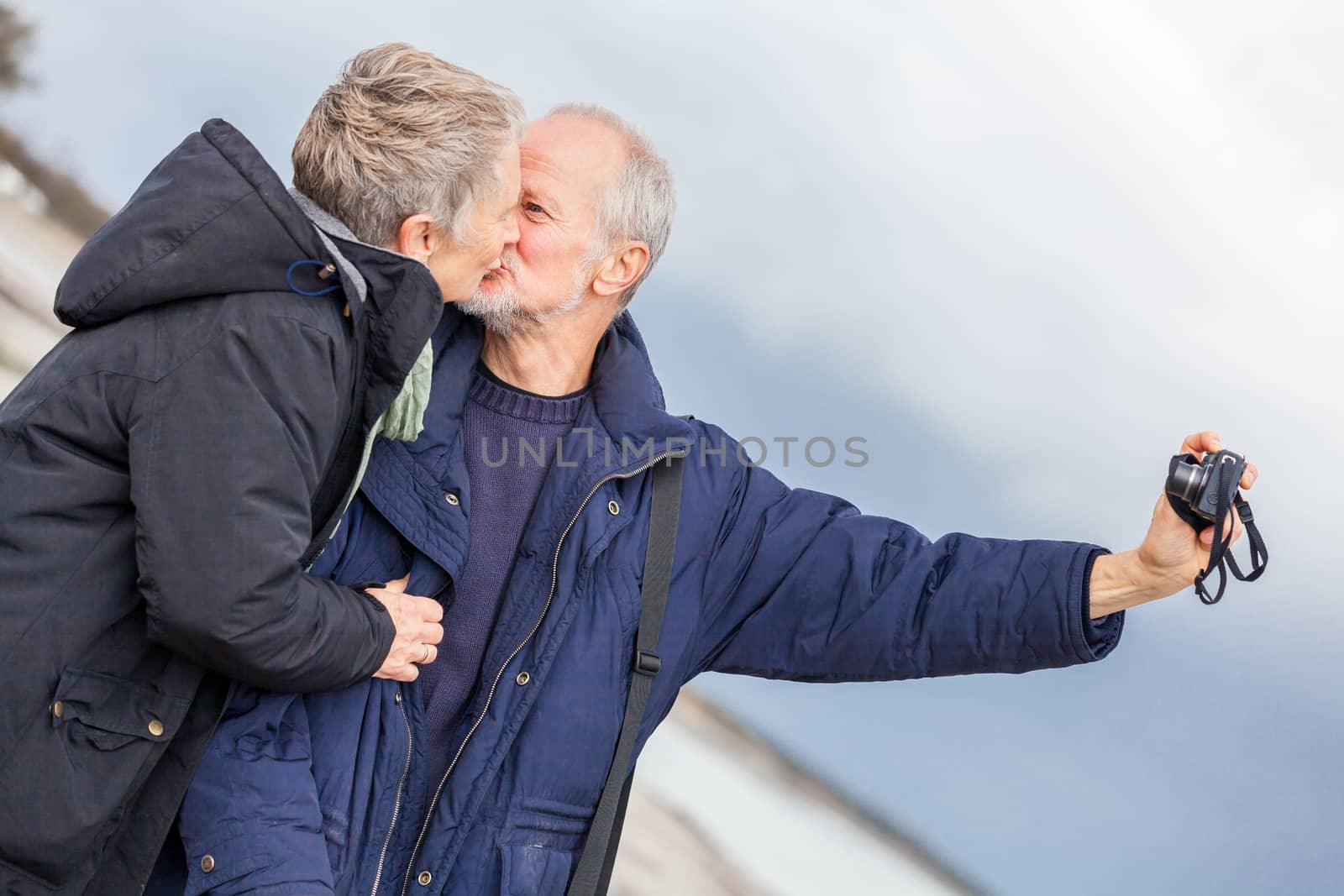 Elderly couple taking a self portrait on a compact digital camera posing in the open air and sunshine with their heads close together smiling at the lens