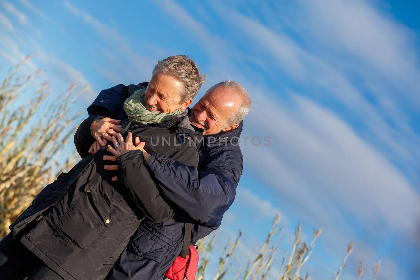 Attractive elderly couple in warm clothing standing clue together with outstretched arms, closed eyes and laughing smile against a blue sky embracing and celebrating the sun