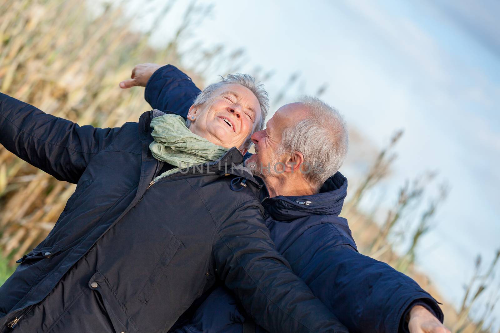 Attractive elderly couple in warm clothing standing clue together with outstretched arms, closed eyes and laughing smile against a blue sky embracing and celebrating the sun