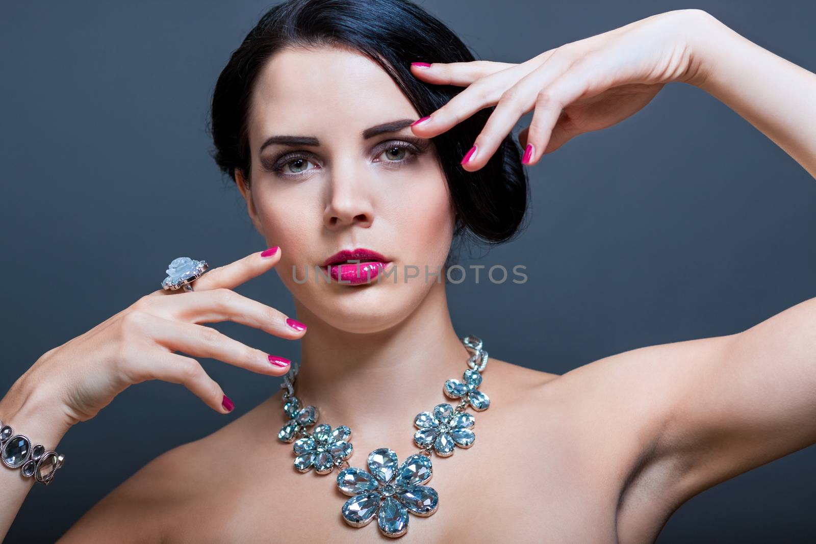 Beautiful sophisticated dark haired woman wearing elegant showy gemstone jewellery posing with bare shoulders and her hand raised gracefully to her downcast eyes with a serene expression