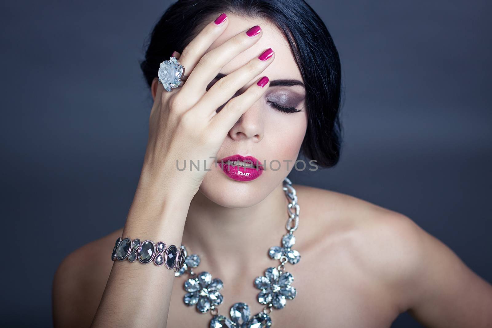 Beautiful sophisticated dark haired woman wearing elegant showy gemstone jewellery posing with bare shoulders and her hand raised gracefully to her downcast eyes with a serene expression