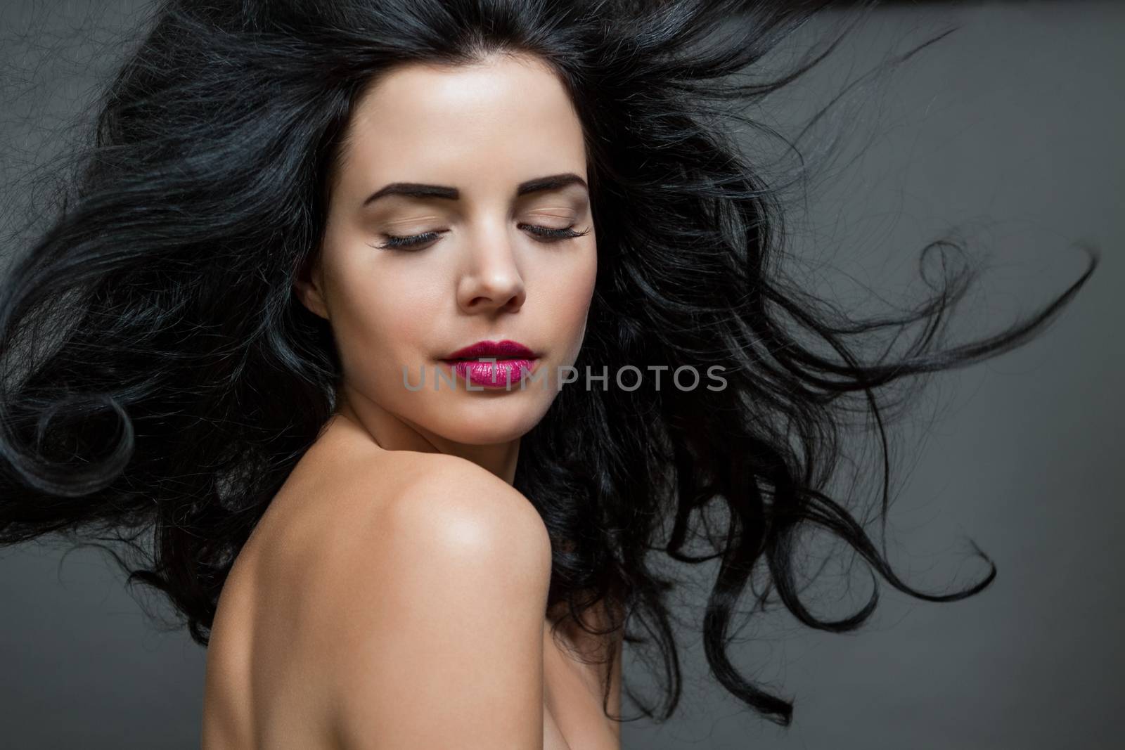 Atmospheric studio portrait of a beautiful black haired woman with a gentle serene expression gazing over her naked shoulder directly at the camera, close up head and shoulders against black