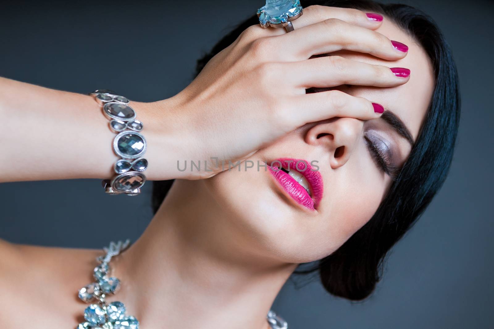 Beautiful sophisticated dark haired woman wearing elegant showy gemstone jewellery posing with bare shoulders and her hand raised gracefully to her downcast eyes with a serene expression