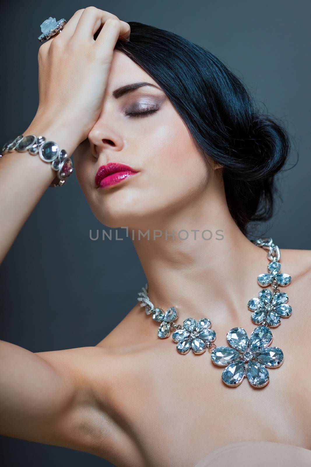 Beautiful sophisticated dark haired woman wearing elegant showy gemstone jewellery posing with bare shoulders and her hand raised gracefully to her downcast eyes with a serene expression