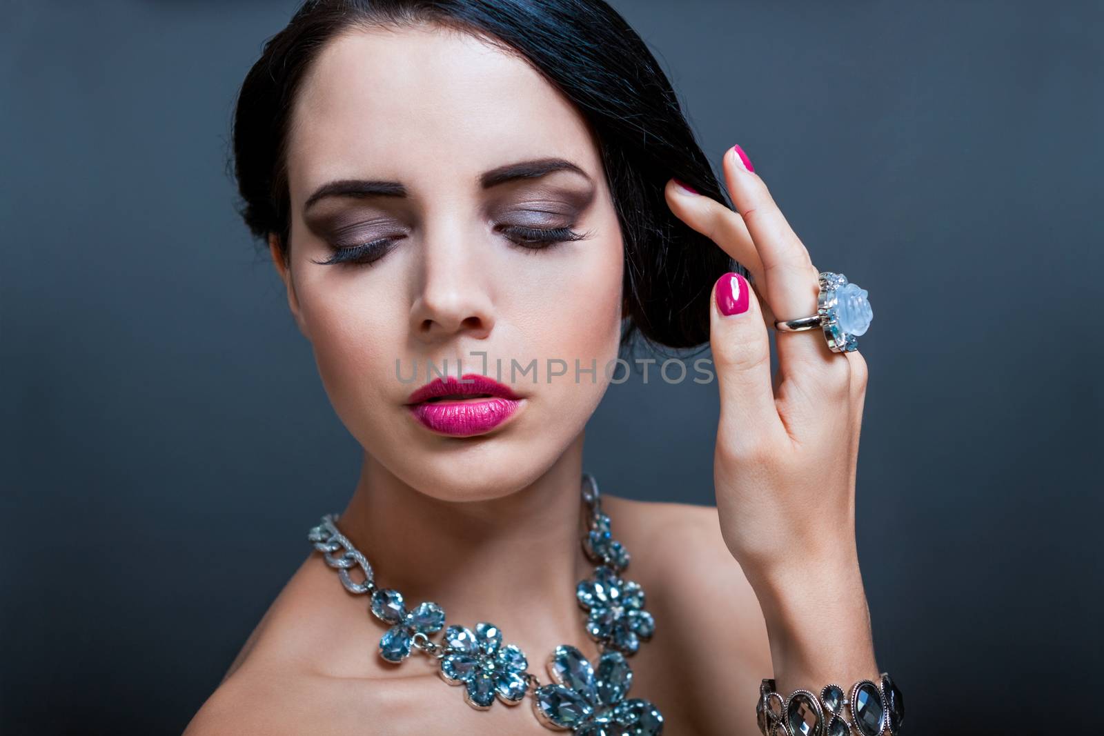 Beautiful sophisticated dark haired woman wearing elegant showy gemstone jewellery posing with bare shoulders and her hand raised gracefully to her downcast eyes with a serene expression