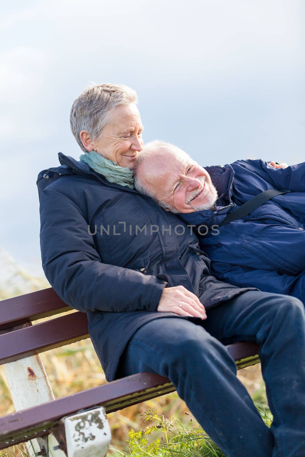 happy senior couple relaxing together in the sunshine on a wooden bench in the countryside with the one reclining full length on the seat with his head on his partners lap