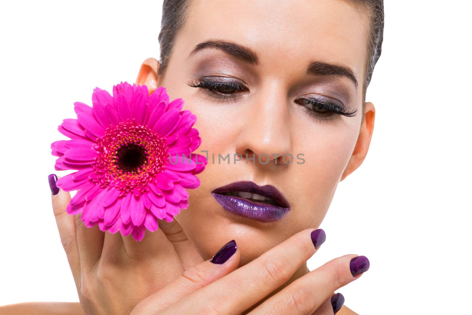 Close up facial portrait of a beautiful young brunette woman in purple make-up with her hand resting gracefull on her cheek holding a fresh magenta Gerbera daisy, isolated on white