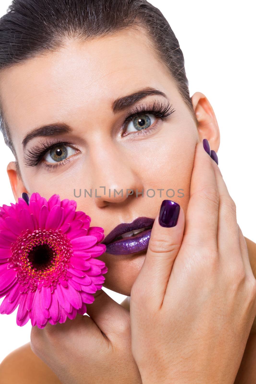 Close up facial portrait of a beautiful young brunette woman in purple make-up with her hand resting gracefull on her cheek holding a fresh magenta Gerbera daisy, isolated on white