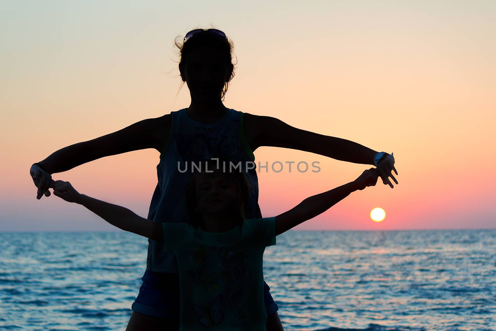 Silhouettes of a young woman and child, standing by the sea , holding hands, on a background of a sunset.