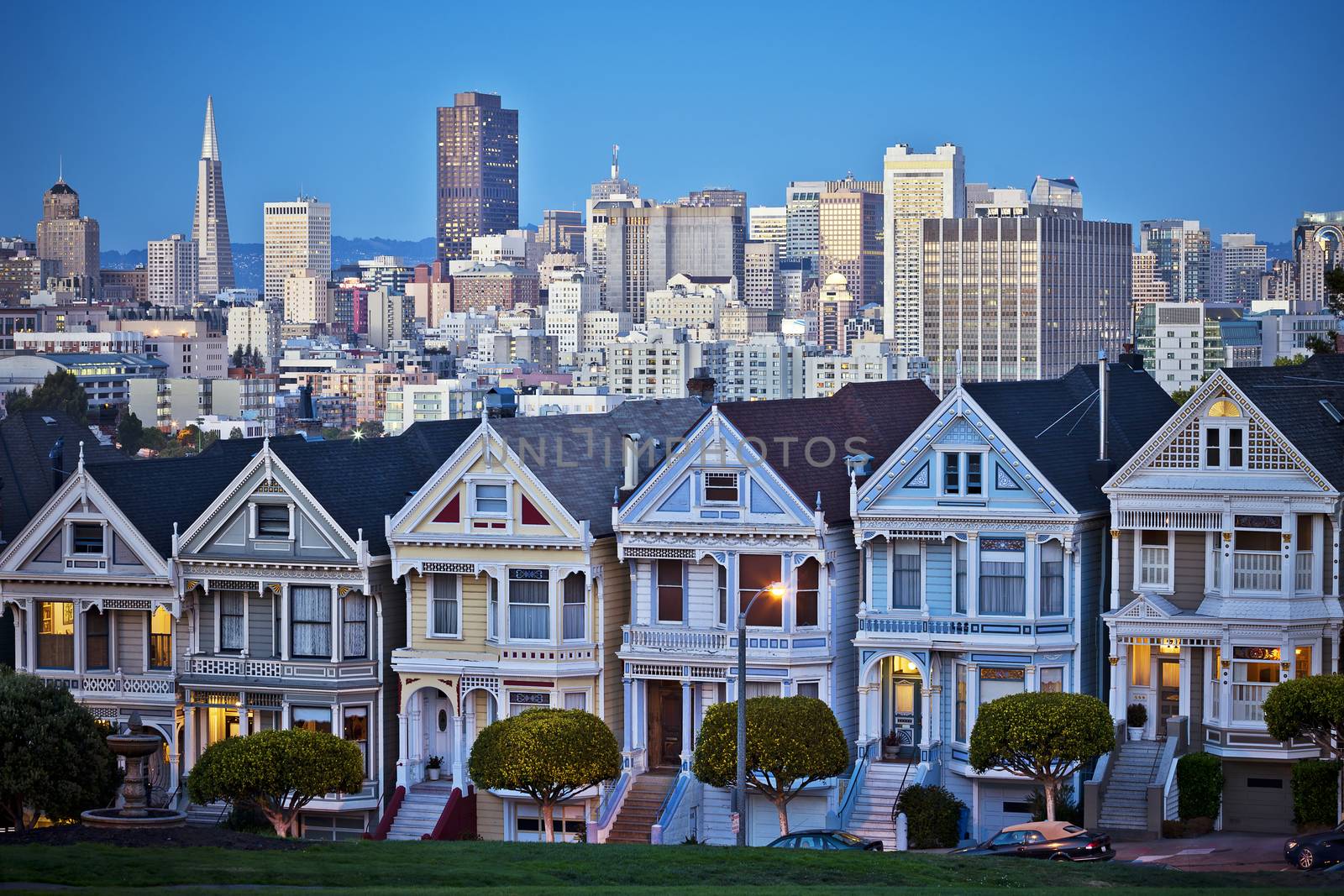 Famous Painted Ladies of San Francisco, California sit glowing amid the backdrop of a sunset and skyscrapers. 
