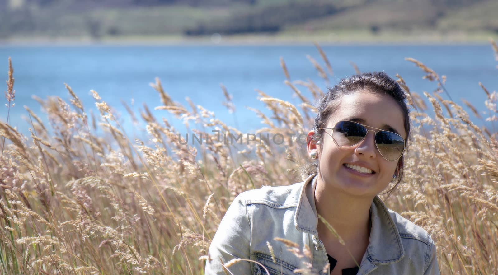 Nice young woman into a golden grass field, she showing happy with a big smile, in the background it can observe a blue lake and the slope of a hill 