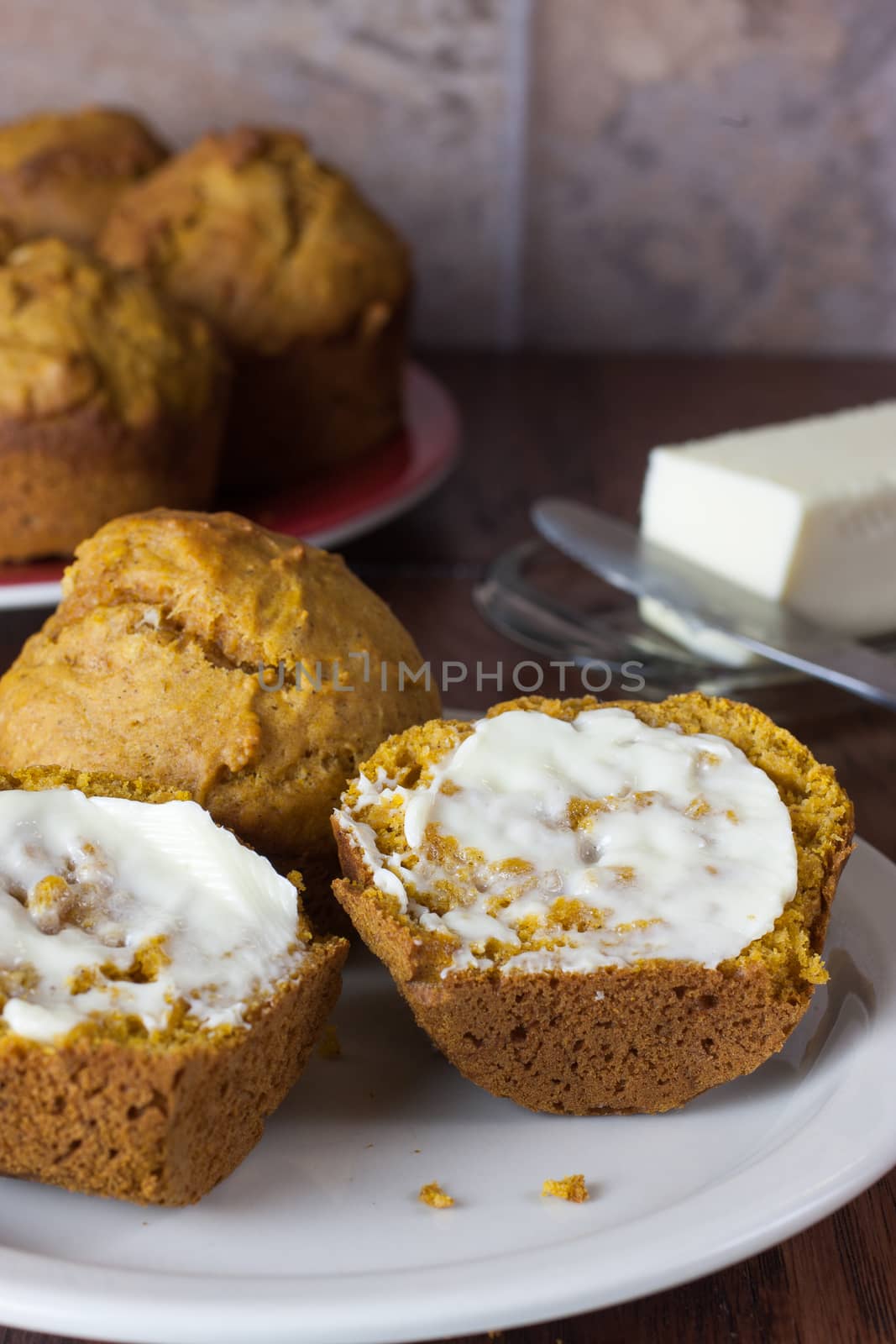 A cut and buttered pumpkin muffin opn a white plate with a plate of muffins in the background.