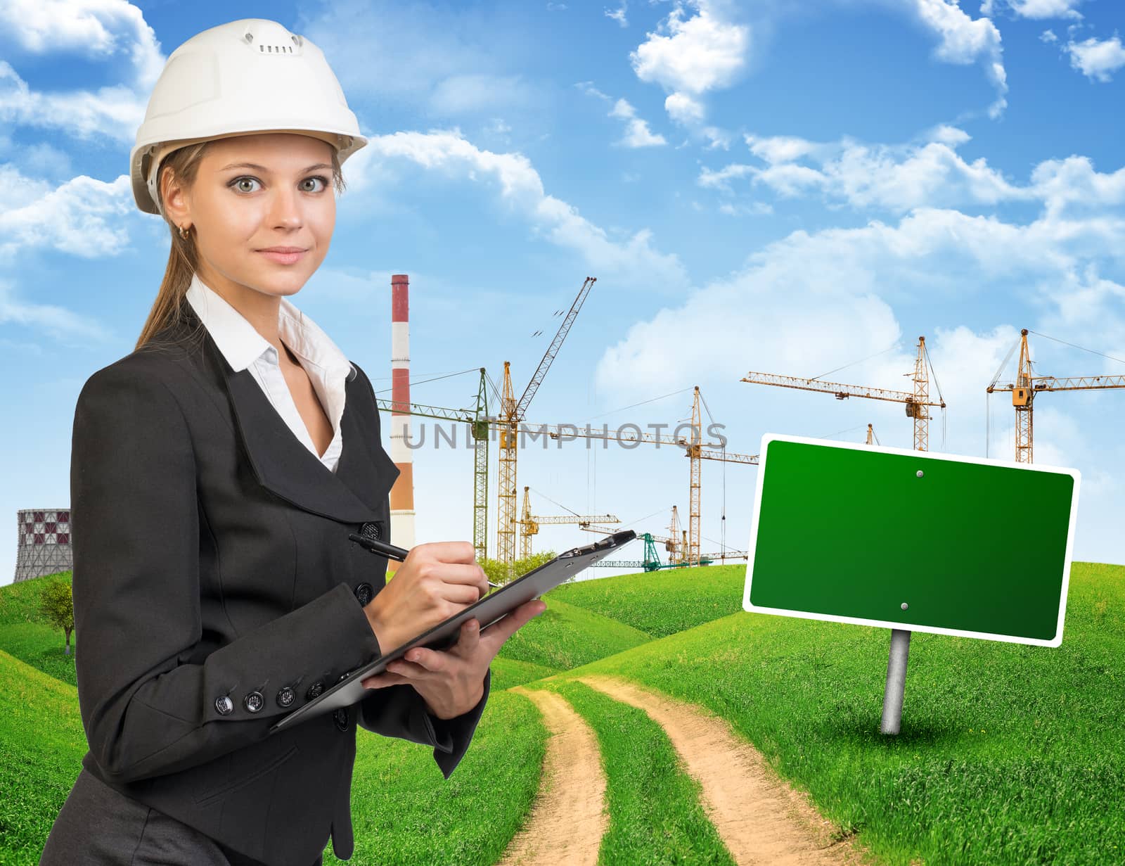 Businesswoman in hard hat standing on the road among green hills, writing on clipboard, looking at camera. Further off blank green billboard out of upright. Cranes and smoke-stacks as backdrop