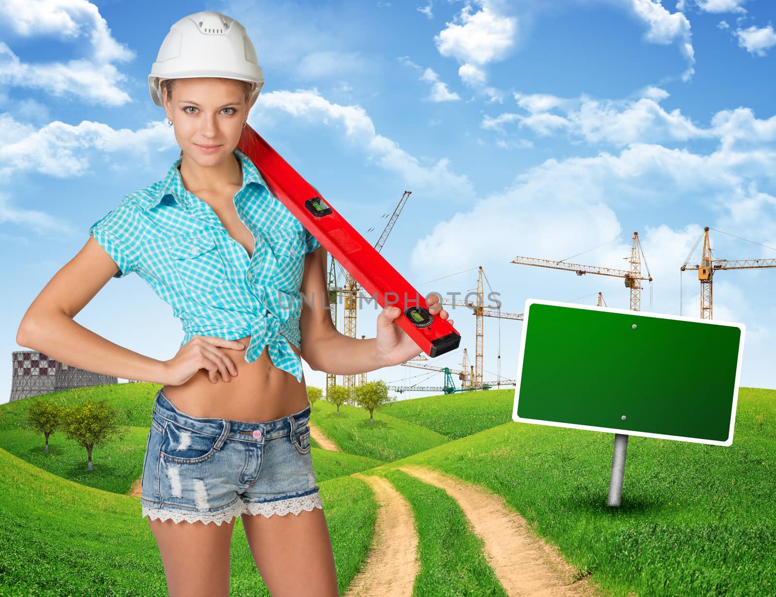 Woman in hard hat, holding large spirit level on her shoulder, standing on the road among green hills, looking at camera, smiling. Further off blank green billboard out of upright. Cranes and smoke-stacks as backdrop