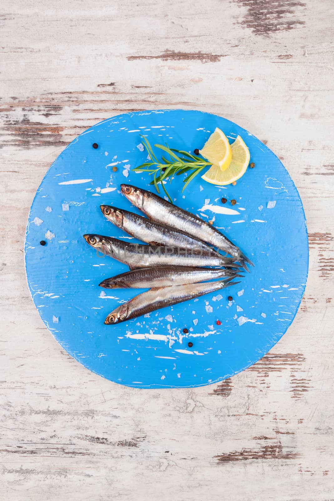Several sardines on blue round kitchen board, on white wooden background, top view. Mediterranean seafood eating.