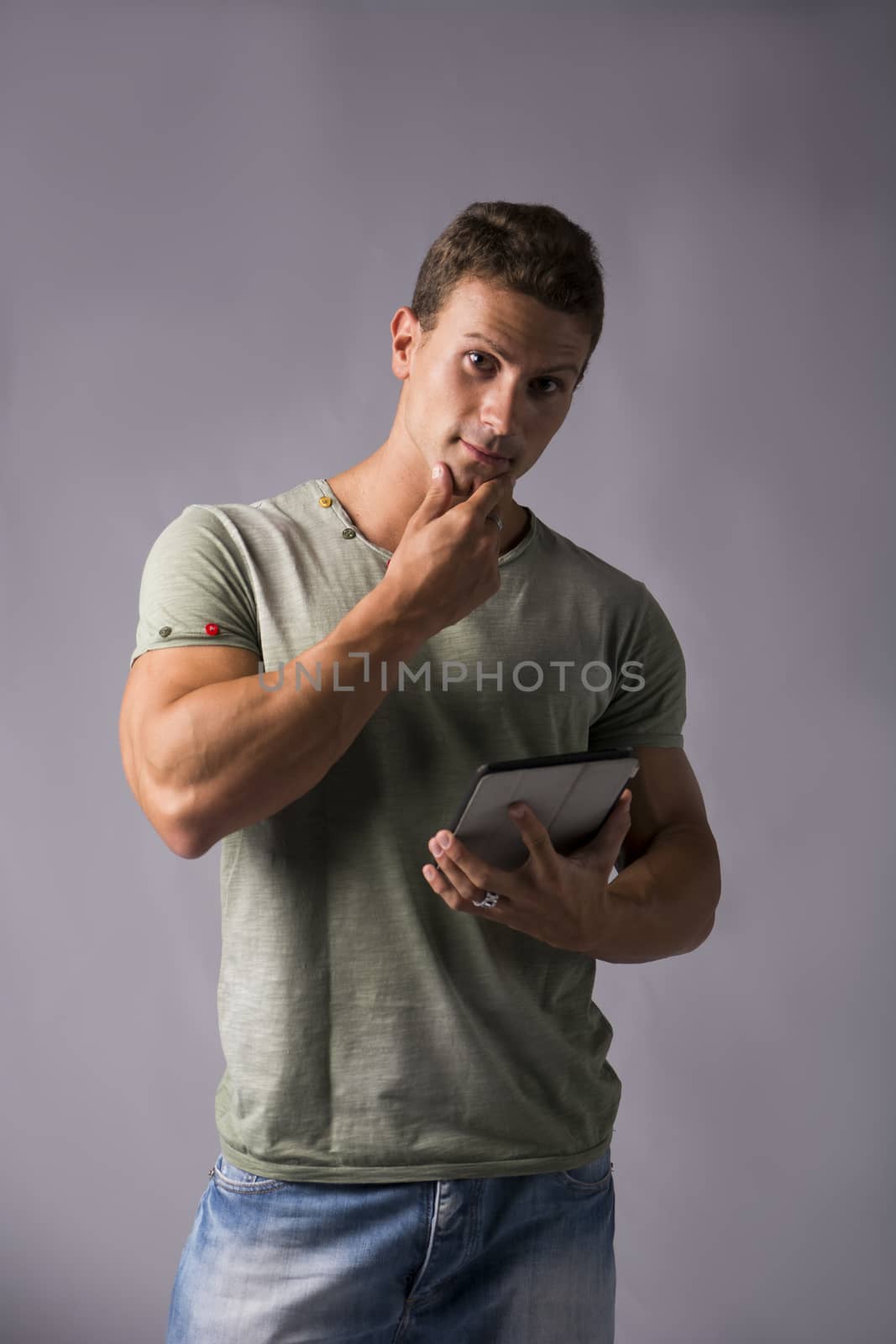 Attractive young man holding ebook reader or tablet computer and holding his chin, standing on grey background