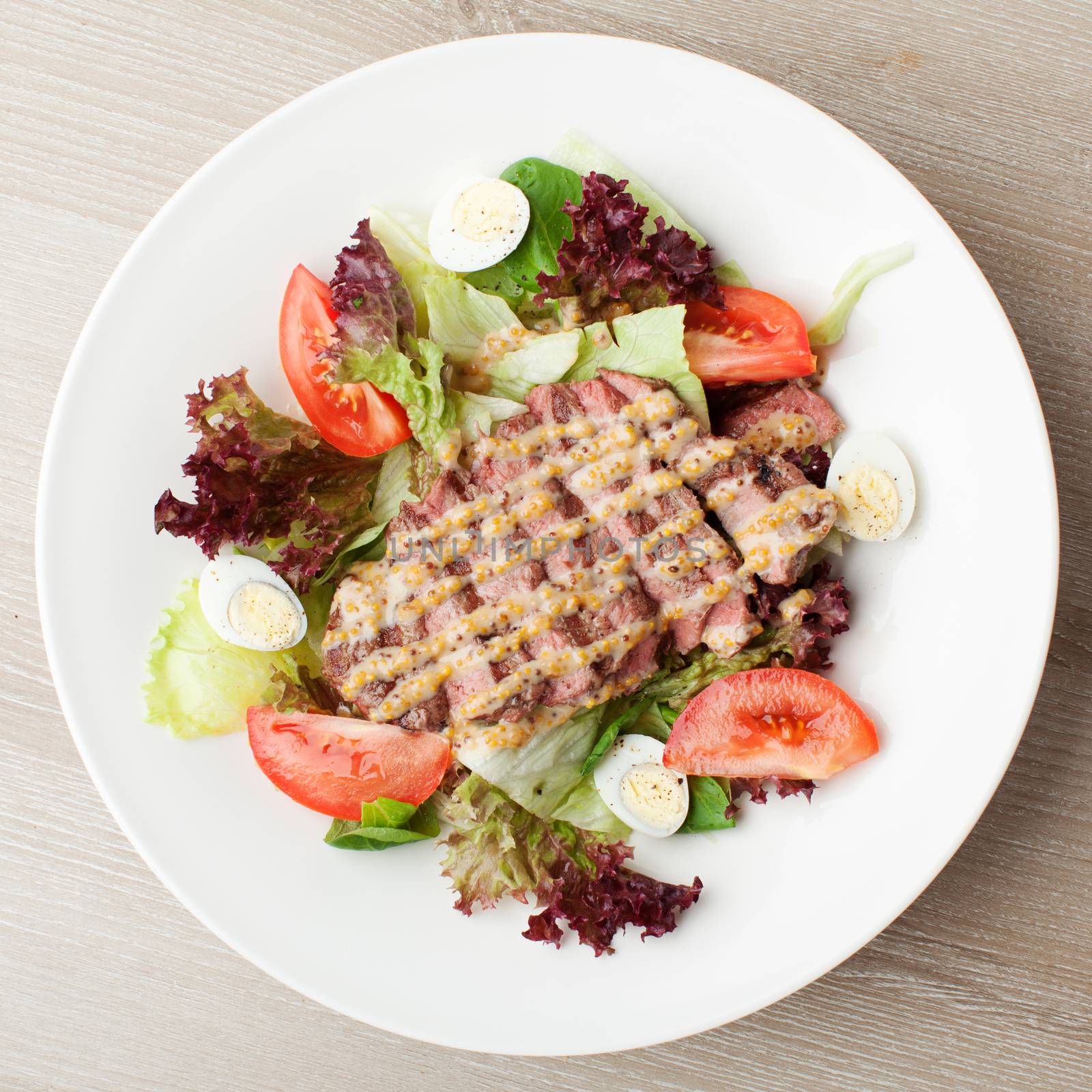 Fresh salad mix with grilled beef, meat cherry tomatoes , boiled quail eggs and  mustard  sauce served on table in white plate