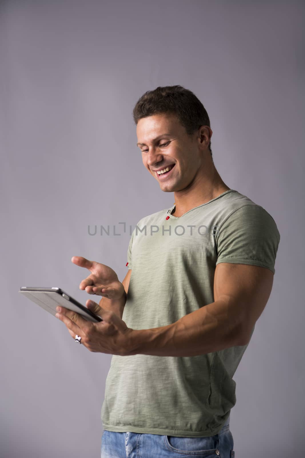 Attractive young man holding ebook reader or tablet PC laughing amused by what he sees, standing on grey background