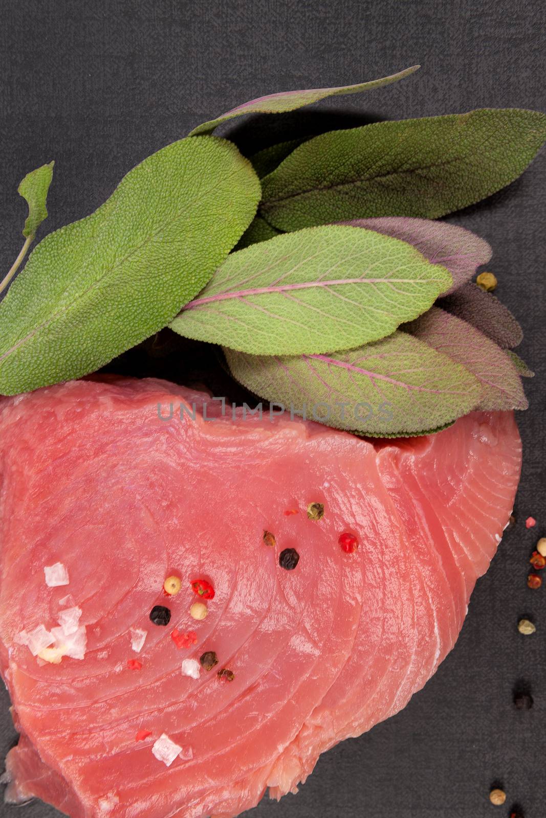Raw fresh tuna steak with fresh sage herbs, peppercorns and salt on black background, top view.