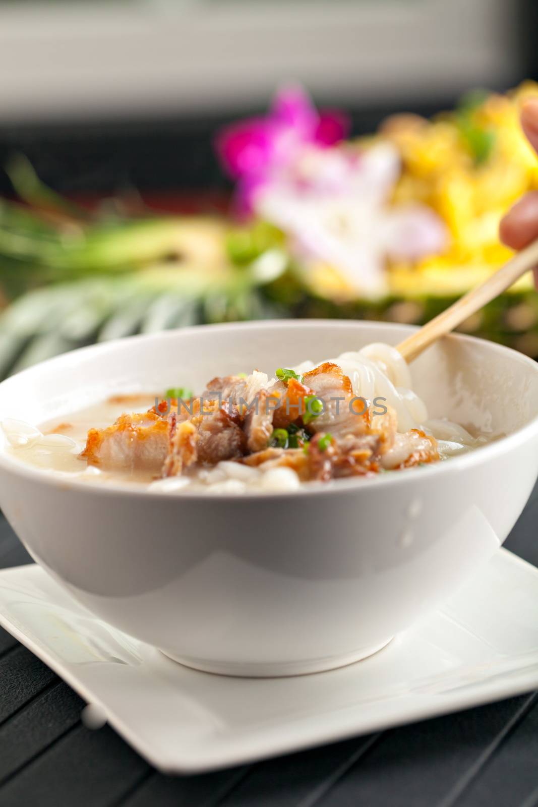 Closeup of a person eating Thai style crispy pork rice noodle soup from a bowl with chopsticks. Pineapple fried rice in the background.