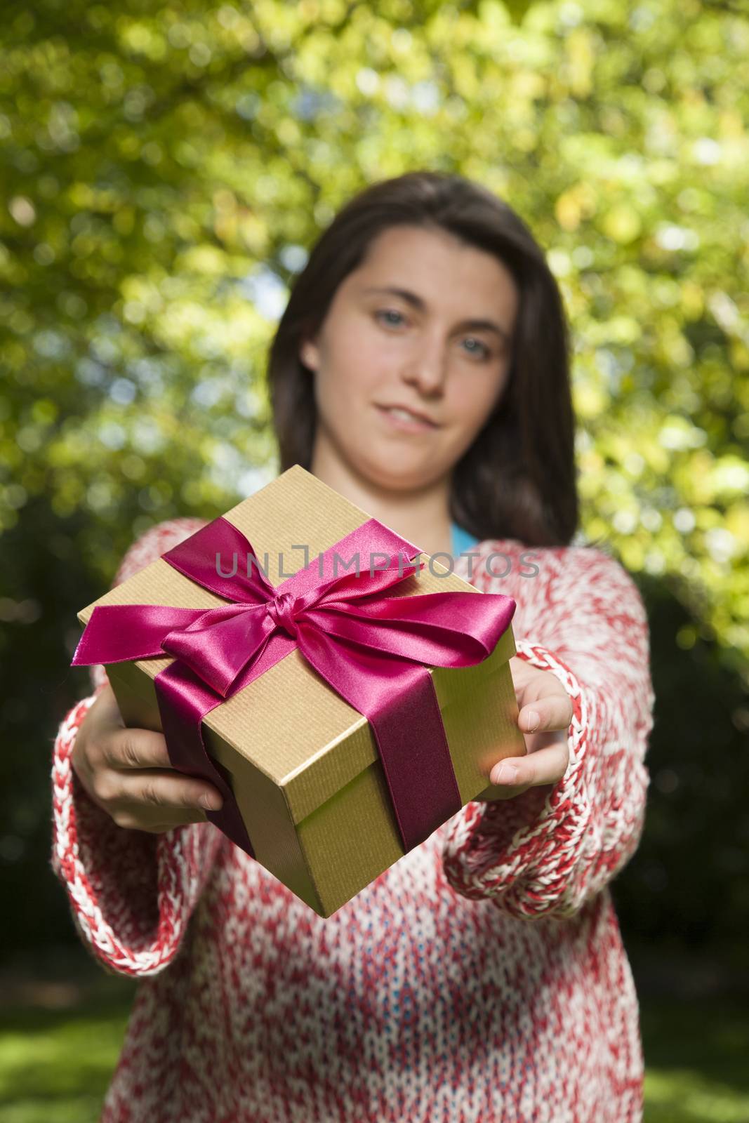 young woman offering golden red gift box over green tree background
