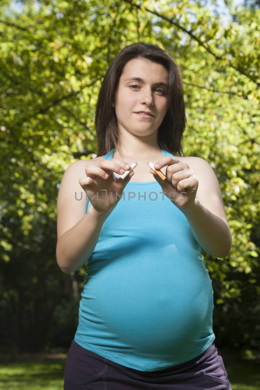 young pregnant woman blue shirt breaking cigarette over green tree background