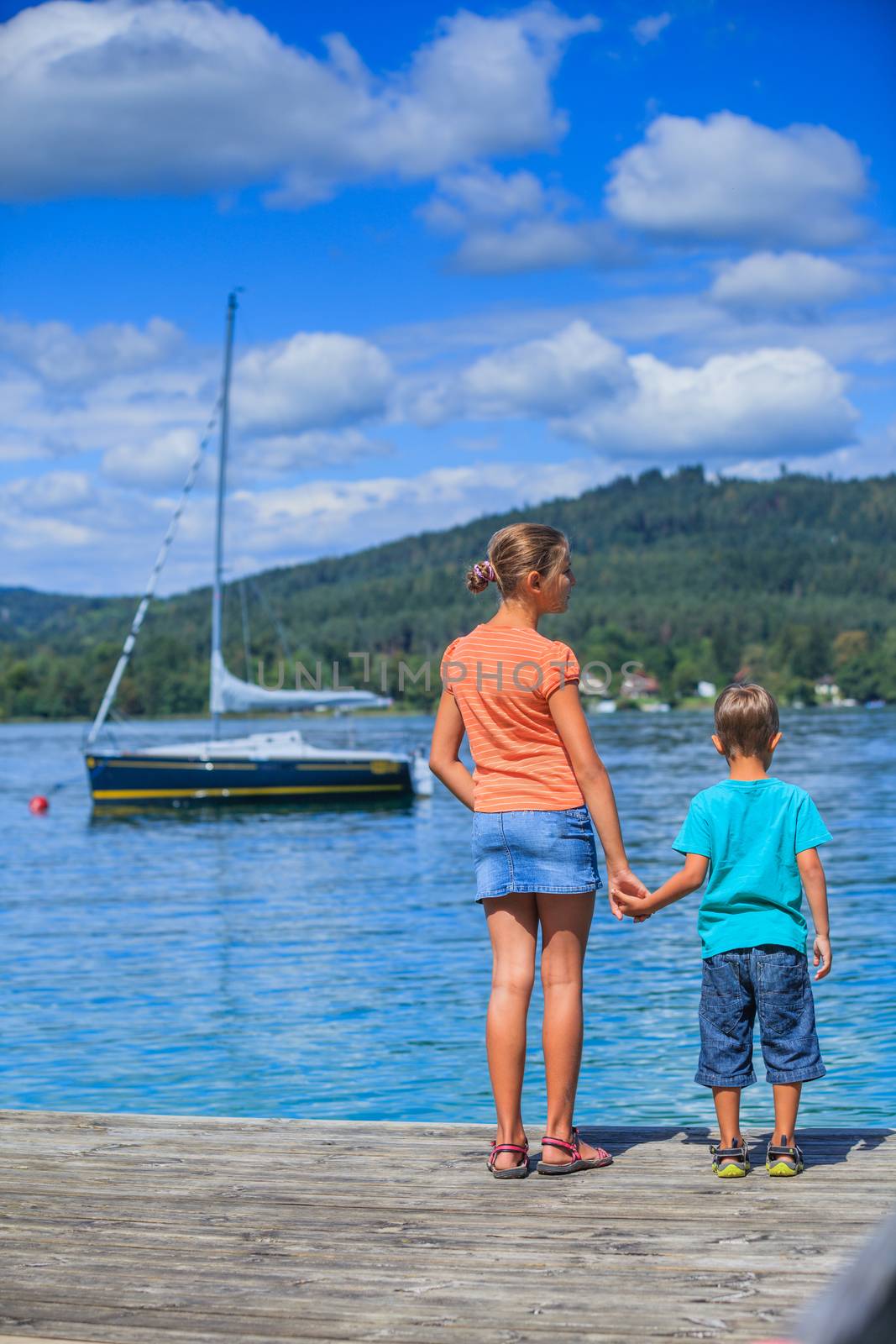 Summer vacation  at the lake - Back view of two happy kids walking on the pier and watching on the yacht