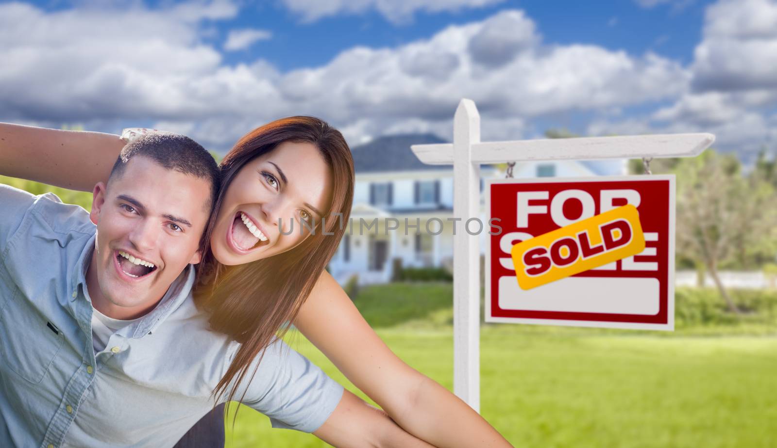 Playful Excited Military Couple In Front of Home with Sold Real Estate Sign.