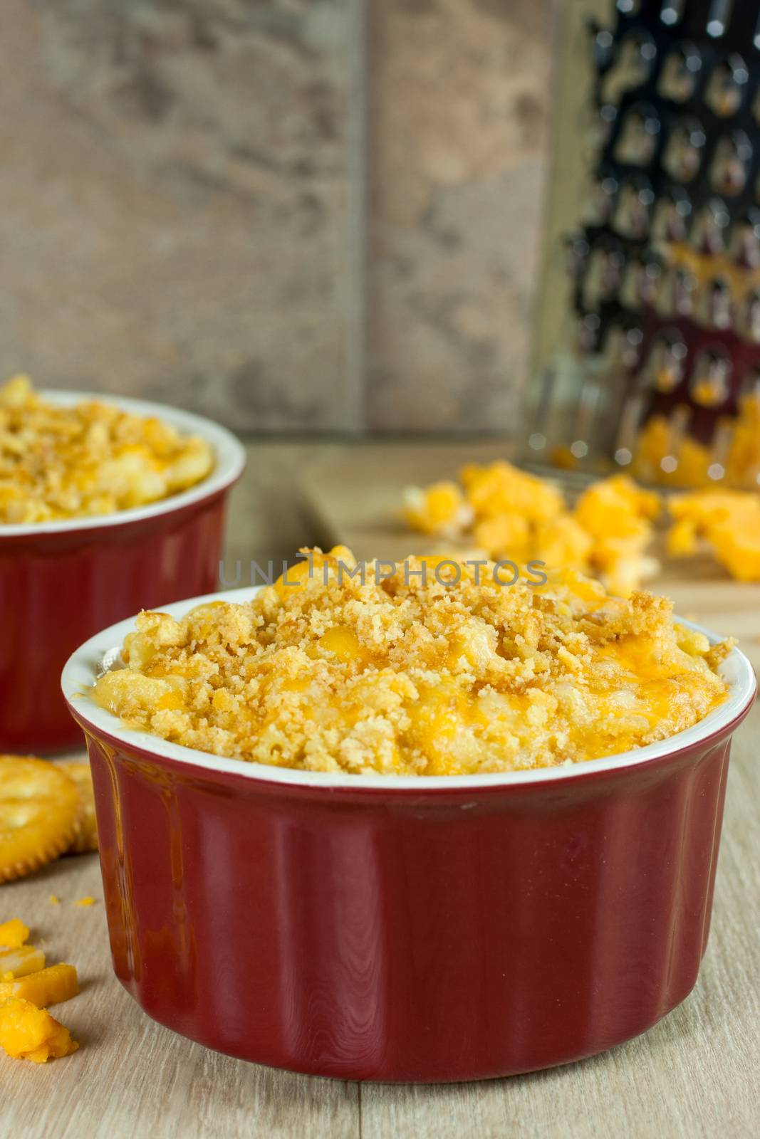 Baked macoroni and cheese topped with a crispy crust in red stone crocks with grated cheese in the background.
