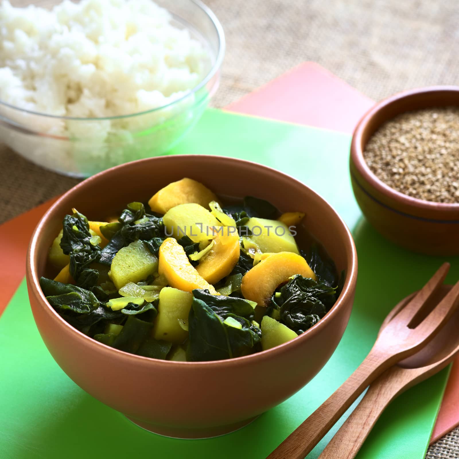 Bowl of spinach, peach and potato curry dish with sesame seeds and cooked rice in the back photographed with natural light (Selective Focus, Focus in the middle of the curry dish)