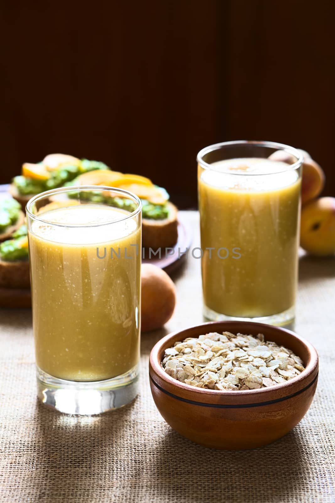 Mango-oatmeal milkshake with a bowl of rolled oats on the side and avocado sandwiches in the back photographed with natural light (Selective Focus, Focus on the front rim of the glass and the front of the oats in the bowl) 