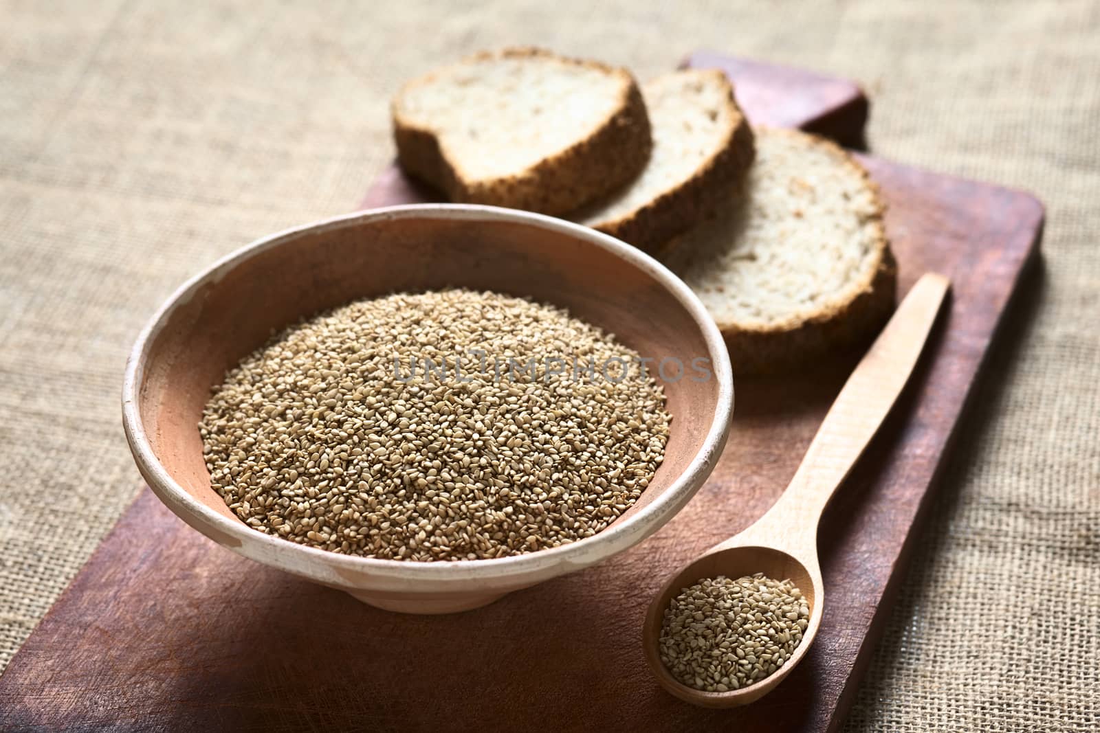 Sesame seeds in bowl and wooden spoon with wholegrain bread in the back photographed with natural light (Selective Focus, Focus one third into the seeds)