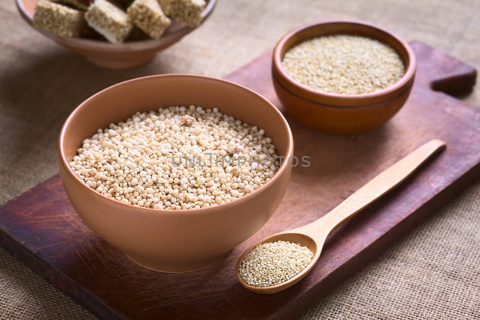 Popped white quinoa (lat. Chenopodium quinoa) cereal in bowl with raw quinoa seeds and quinoa cereal bars in the back photographed with natural light (Selective Focus, Focus one third into the quinoa cereal)