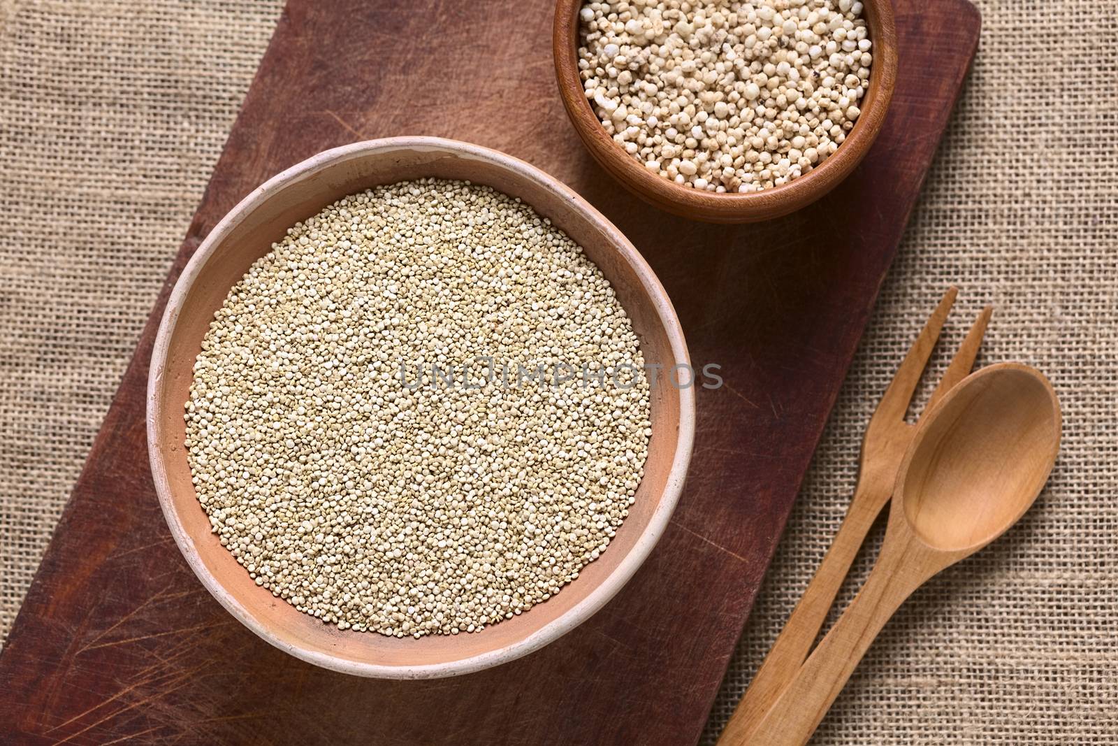 Overhead shot of raw white quinoa (lat. Chenopodium quinoa) grain seeds in bowl with popped quinoa cereal on wooden board photographed with natural light