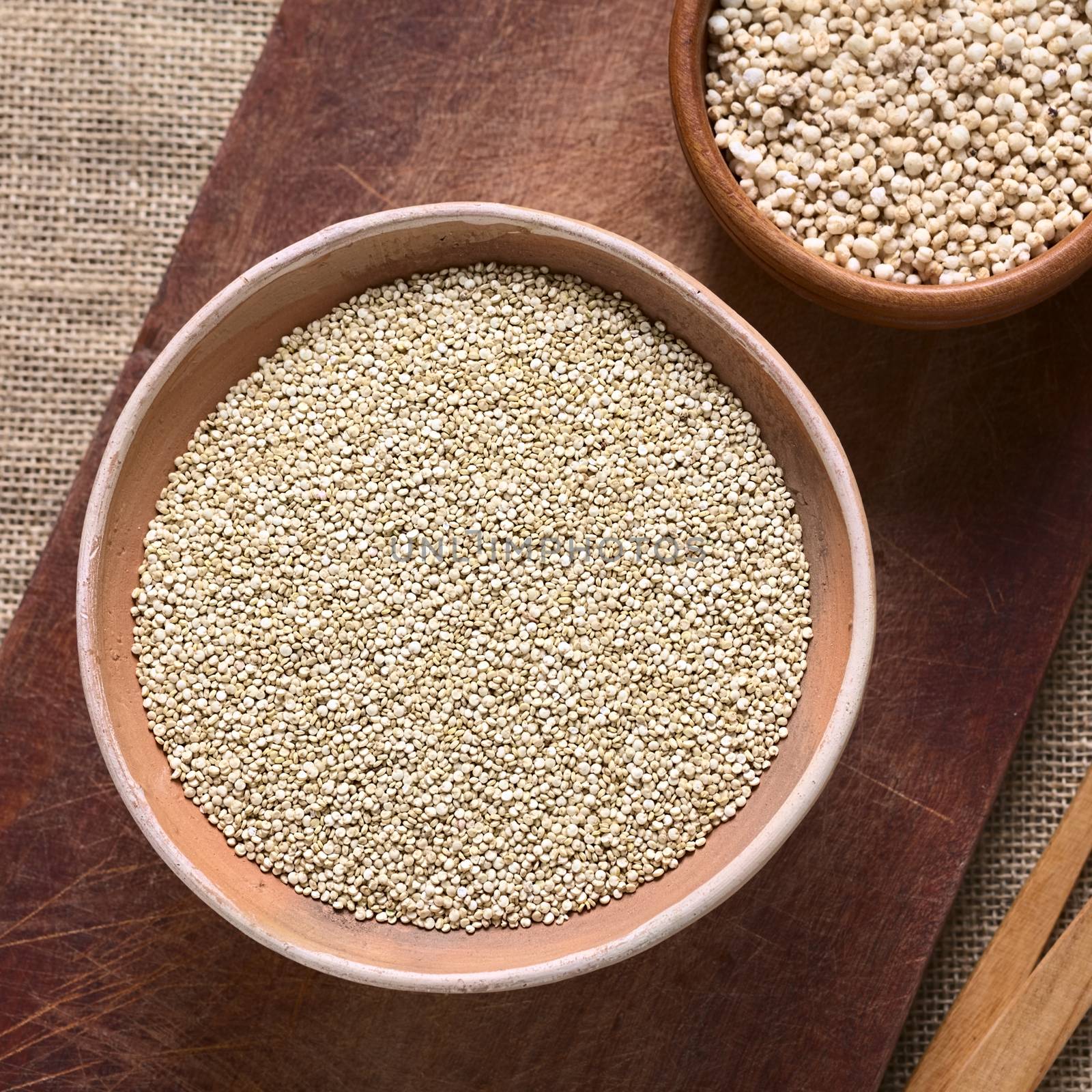 Overhead shot of raw white quinoa (lat. Chenopodium quinoa) grain seeds in bowl with popped quinoa cereal on wooden board photographed with natural light