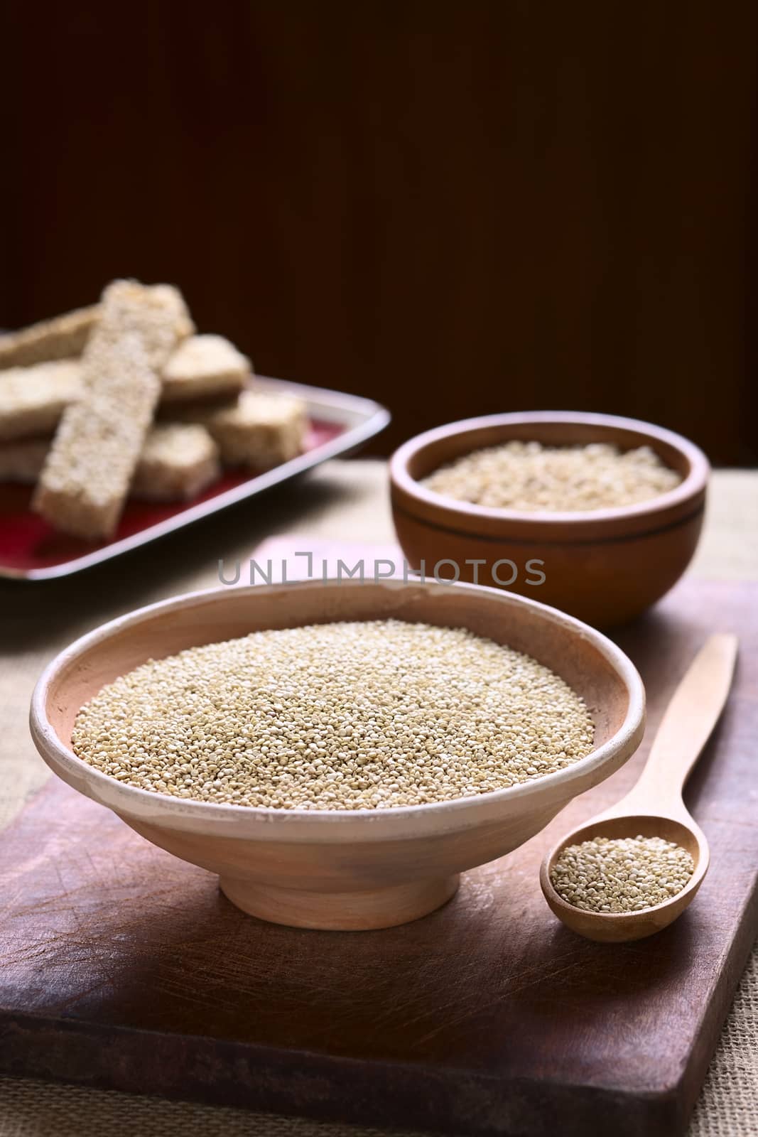 Raw white quinoa (lat. Chenopodium quinoa) grain seeds in bowl with quinoa cereal bars in the back on wooden board photographed with natural light (Selective Focus, Focus one third into the raw quinoa seeds)