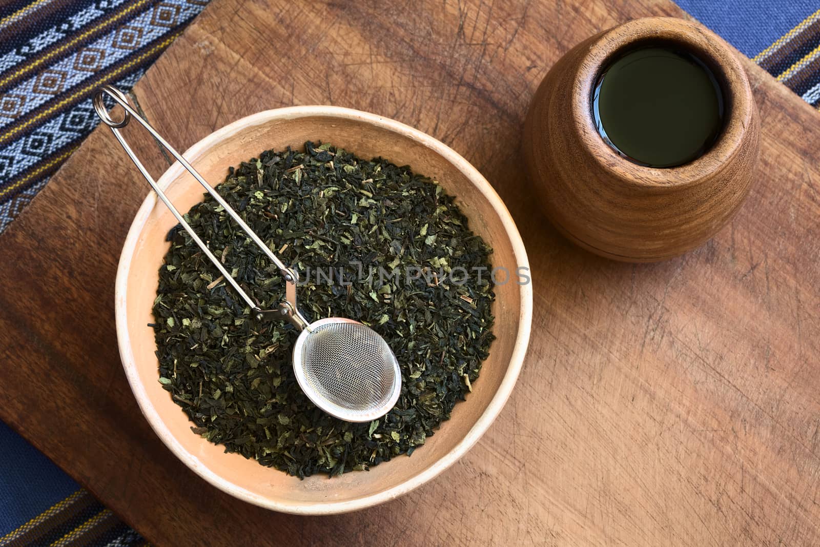 Overhead shot of dried green tea leaves in bowl with a tea strainer on top and wooden tea cup on the side photographed with natural light