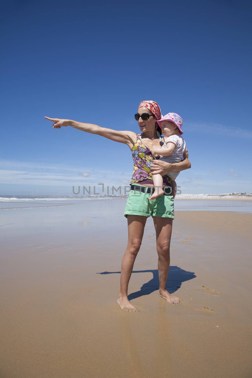 happy woman with sunglasses green shorts headscarf and baby pink hat in arms at beach Conil Cadiz Spain