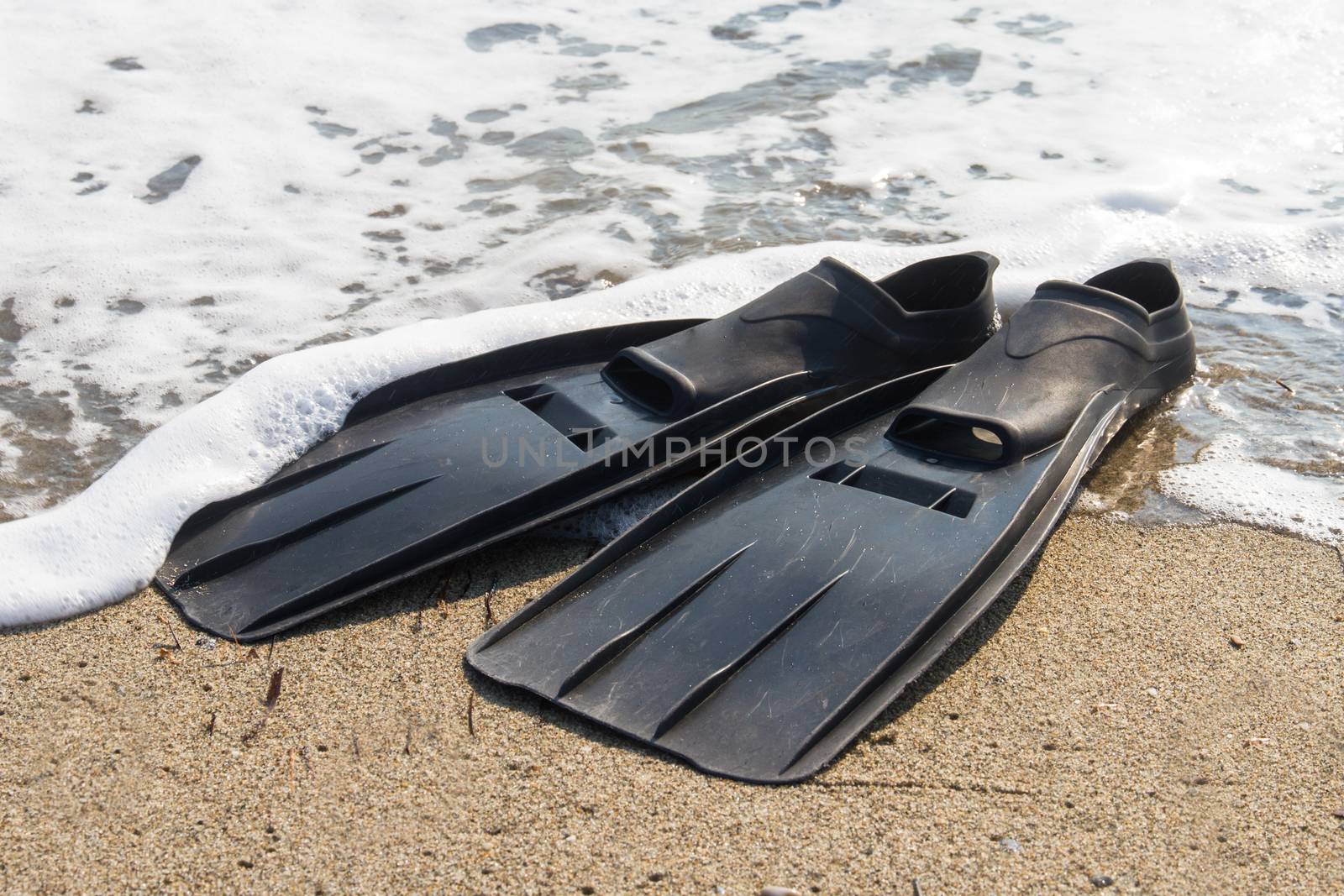 Black plastic flippers on sea sand and wavy sea with foam.